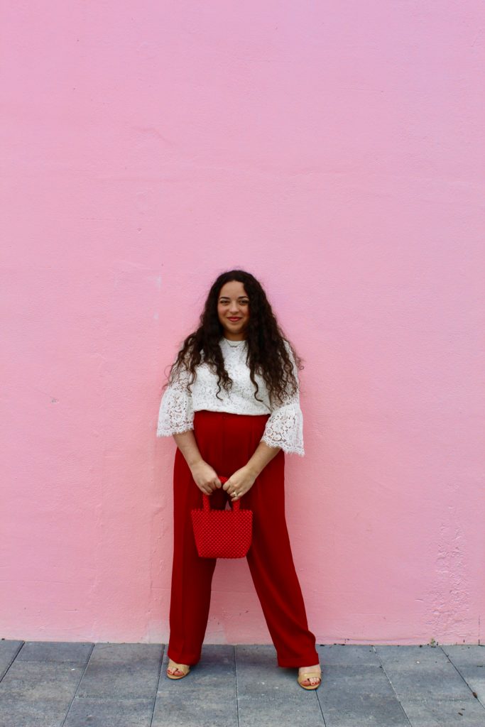 woman standing in front of a pink wall wearing white lace blouse, red pants, pink Zara bag, nude heels